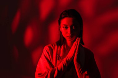 Kimono pelerinli genç Asyalı kadın dua eden elleri kırmızı ışıkta, koyu arka planda duruyor.