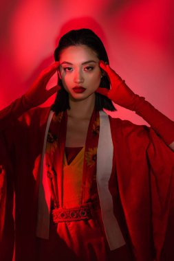 Kimono pelerinli ve eldivenli genç Asyalı kadın soyut arka planda kırmızı ışıkla ellerinin yanında poz veriyor.
