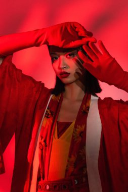 Zarif kimono pelerinli genç Asyalı kadın ve soyut arka planda kırmızı tonda poz veren eldivenler.