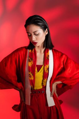 Eşarplı esmer Asyalı kadın çiçek dekoru ve kimono pelerinli soyut kırmızı arka planda elleri kalçasında poz veren