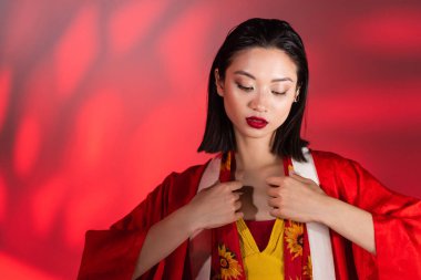 Kimono pelerinli genç Asyalı kadın kırmızı gradyan ile soyut arka planda çiçek desenli atkıya dokunuyor. 