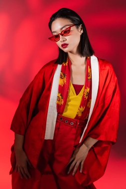 fashionable asian woman in kimono cape and sunglasses posing on red background with shadow clipart