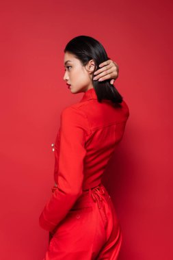 side view of brunette asian woman in stylish suit fixing hair while looking away on red background clipart