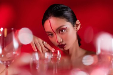 Yaratıcı makyajlı genç Asyalı kadın kırmızı arka planda bulanık gözlüklü kameraya bakıyor.