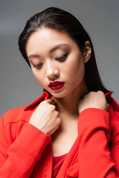 portrait of brunette asian woman with makeup touching collar of red jacket isolated on grey