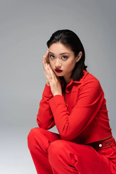 穿着红色雅致服装的时髦的亚洲女人坐在后腿上 双手靠近脸 灰色孤立 — 图库照片