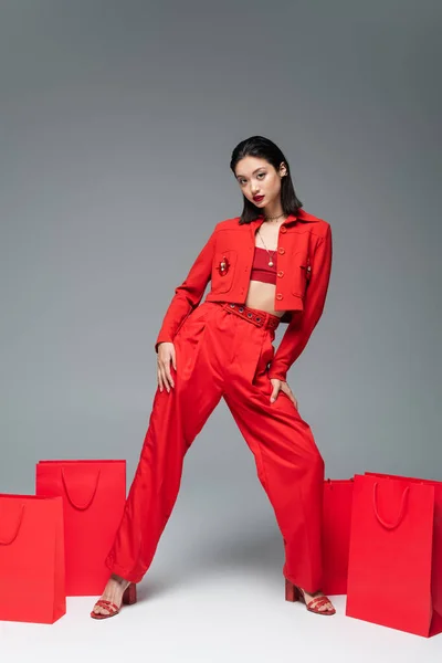 全长的亚洲模特 穿着红色夹克和裤子 在灰色背景的购物袋附近摆姿势 — 图库照片