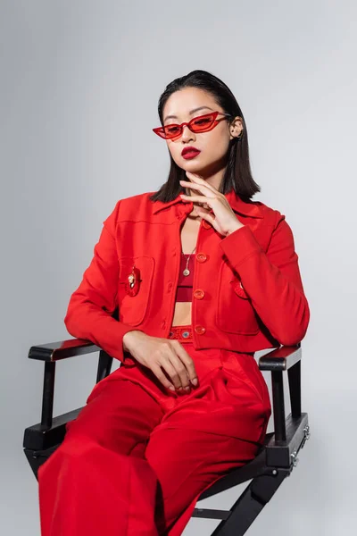 黑发亚洲女人 戴着红色太阳镜 穿着雅致的衣服 坐在椅子上 摸着灰色的下巴 — 图库照片