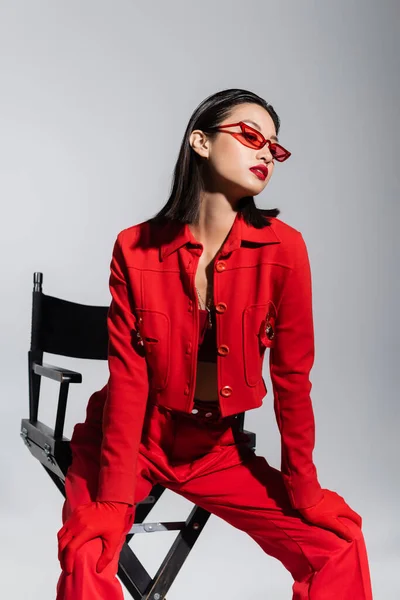 雅致的亚洲模特 身穿红色夹克 戴着太阳镜 躺在椅子上 与灰色隔离 — 图库照片