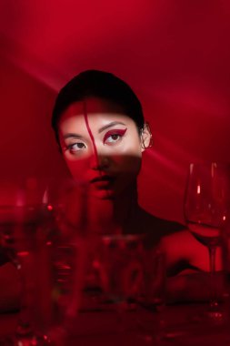 young asian woman with artistic makeup looking away in light near blurred glasses on dark red background clipart