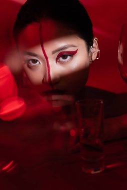 asian woman with creative visage and ear cuff looking at camera in light near blurred glasses on dark red background clipart