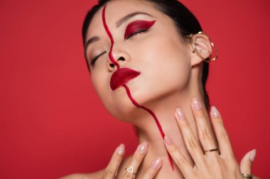 seductive asian woman with ear cuff and creative makeup posing with closed eyes and hands near neck isolated on red clipart