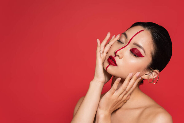seductive asian woman with naked shoulders posing in artistic makeup and stylish ear cuff isolated on red