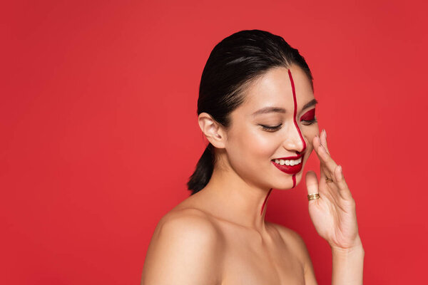 young asian woman with naked shoulders and artistic visage smiling with closed eyes isolated on red