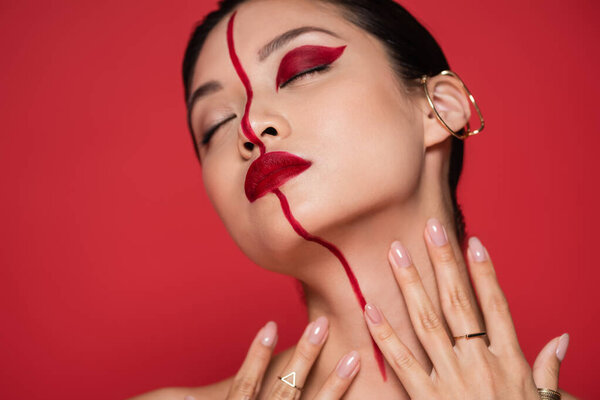seductive asian woman with ear cuff and creative makeup posing with closed eyes and hands near neck isolated on red