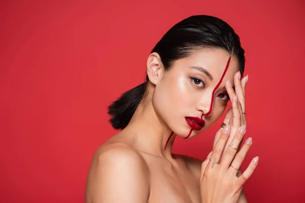 stock image sexy asian woman with naked shoulders touching face with artistic makeup and looking at camera isolated on red