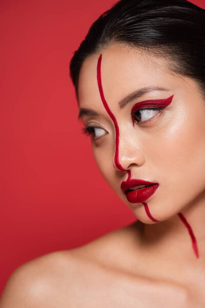 portrait of sensual asian woman with bare shoulder and creative makeup looking away isolated on red