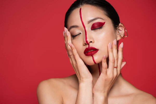 seductive asian woman with bare shoulders and artistic visage touching face and posing with closed eyes isolated on red