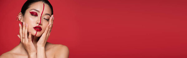 seductive asian woman with bare shoulders touching perfect face with creative makeup isolated on red, banner