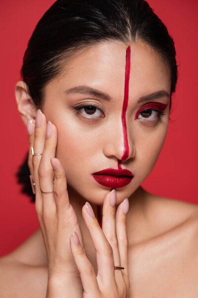 portrait of asian woman touching perfect face with creative makeup and looking at camera isolated on red