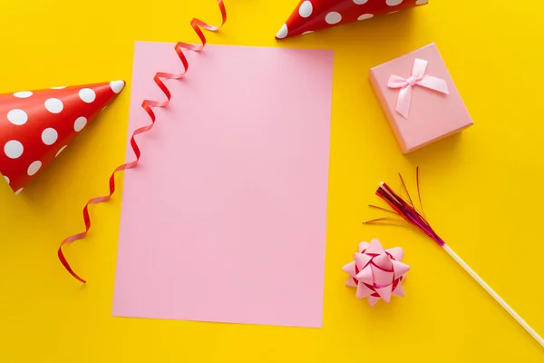 Top view of empty greeting card near dotted party caps and gift box on yellow background