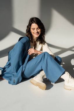 Mavi takım elbiseli ve gri arka planda ışıklandırmalı deri çizmeli neşeli kadının yüksek açılı görüntüsü