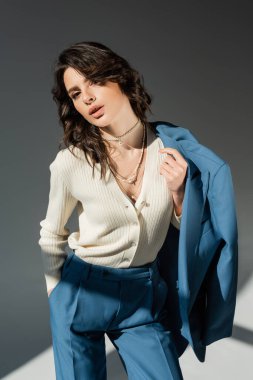 Beyaz kazaklı, kolyeli, ceketli, şık bir kadın gri arka planda mavi pantolonlu.