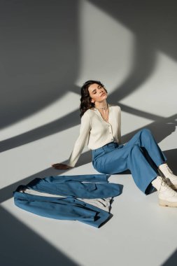 high angle view of brunette woman with closed eyes sitting near blue blazer on grey background with lighting and shadows clipart