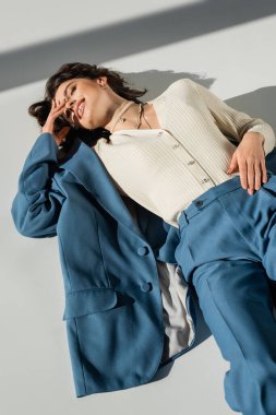 Mavi ve modaya uygun giyinmiş, gri arka planda ışık ve gölgelerle uzanan gülümseyen kadının yüksek açılı görüntüsü 