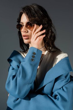 Mavi ceketli esmer kadın gri renkli güneş gözlüklerine dokunuyor.