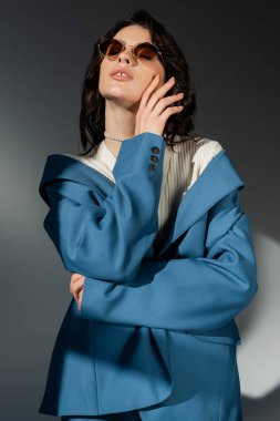 Şehvetli esmer kadın mavi ceketli ve şık güneş gözlüklü gri arkaplana dokunuyor.