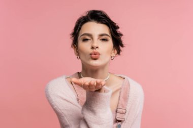 pretty brunette woman in cozy cardigan sending air kiss and looking at camera isolated on pink clipart