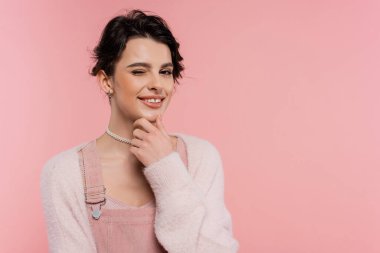 tricky and cheerful woman in fluffy cardigan touching chin and winking at camera isolated on pink clipart