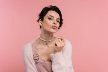 İnci kolyeli şık esmer kadın dudaklarını büzüyor ve pembe kameraya bakıyor.