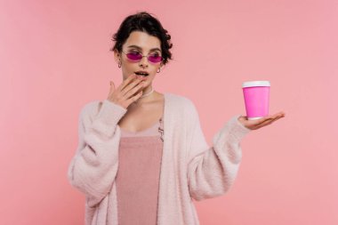 Şık güneş gözlüklü düşünceli bir kadın, ağzının yanında el ele tutuşup pembe üzerine izole edilmiş bir içecek izliyor.