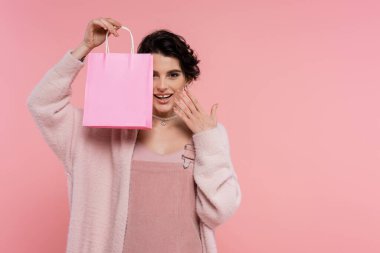 Yumuşak hırka giymiş mutlu bir kadın alışveriş torbası gösteriyor ve pembe üzerinde izole edilmiş yüz yakınında el ele tutuşuyor.