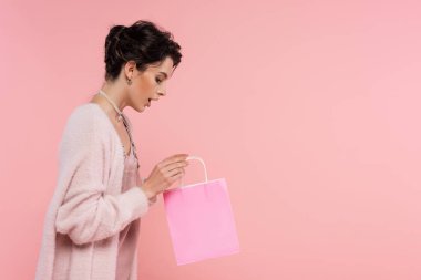Pembe üzerine izole edilmiş alışveriş çantasına bakan kalın hırkalı esmer kadının yan görüntüsü.