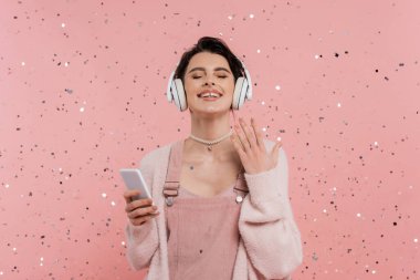 Gözleri kapalı neşeli bir kadın cep telefonu tutuyor ve pembe konfeti yanındaki kablosuz kulaklıkla müzik dinliyor. 