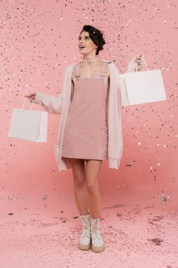 Pembe konfeti altında alışveriş torbalarıyla ayakta duran mutlu ve modaya uygun bir kadın. 