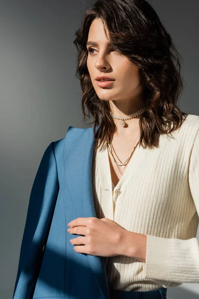 stock image brunette woman in white cardigan and necklaces holding blue blazer and looking away isolated on grey