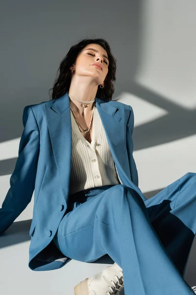 trendy woman in blue suit sitting with closed eyes on grey background with lighting and shadows