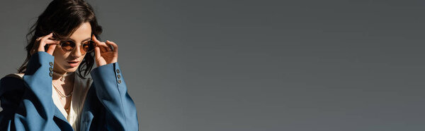 young brunette woman in blue blazer adjusting trendy sunglasses isolated on grey, banner