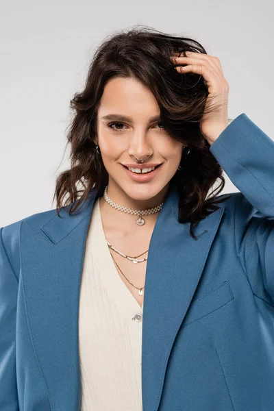 pretty woman with necklaces and piercing posing in blue blazer and smiling at camera isolated on grey