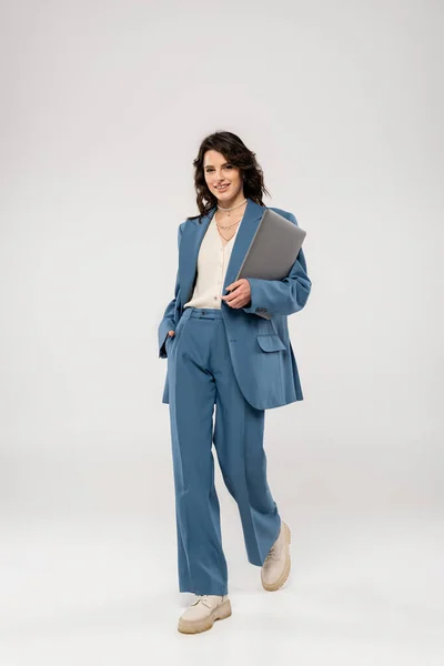 stock image full length of joyful woman in fashionable suit holding hand in pocket while walking with laptop on grey background