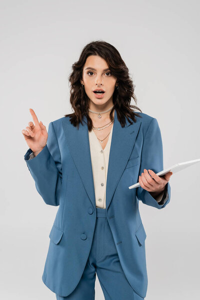 изумленная женщина в синем и модном пиджаке держит цифровой планшет и указывает пальцем, изолированным от серого