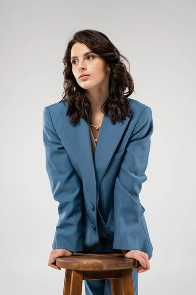 stock image young and stylish woman in blue jacket looking away near wooden stool isolated on grey