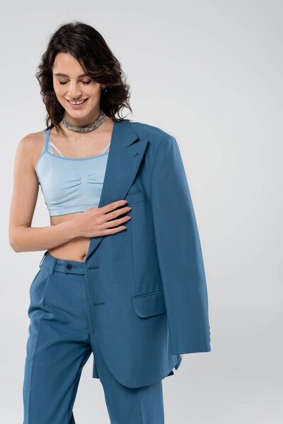 cheerful brunette woman in crop top and pants smiling with closed eyes while holding blazer isolated on grey