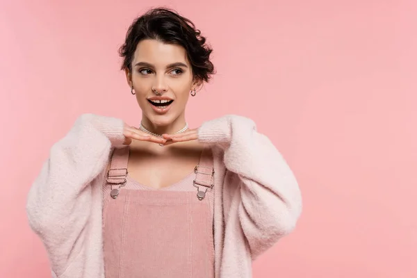 Stock image cheerful woman in strap dress posing with hands under chin and looking away isolated on pink