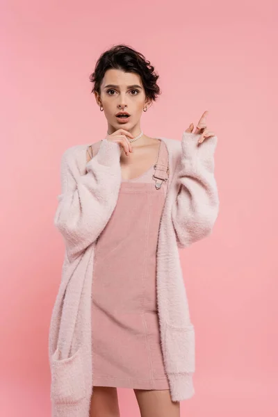 Stock image astonished woman in cozy cardigan touching chin and pointing with finger isolated on pink