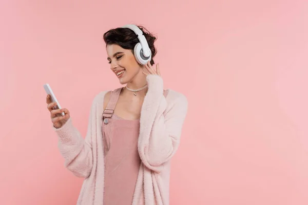 stock image happy brunette woman in wireless headphones looking at mobile phone isolated on pink